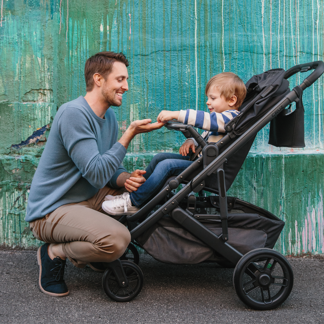 The UPPAbaby Cruz V2 stroller with a toddler eating snacks from the snack tray accessory and handing one to their father that is down on his honkers in front of the stroller. The carry all parent organiser is conveniently attached to the handlebars.