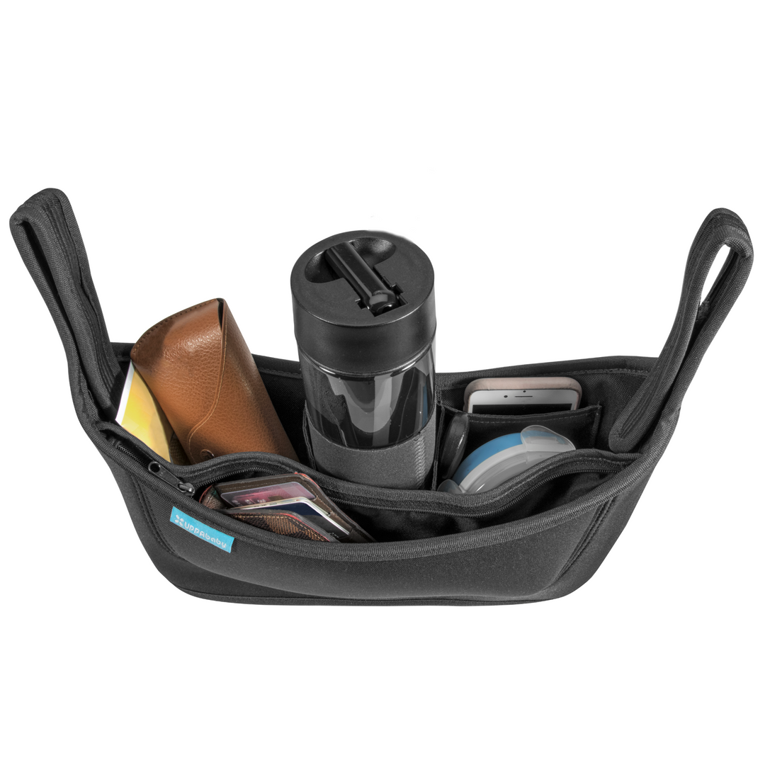 This is an ariel product shot of the stroller organiser to hold parent essentials within reach. It is filled with wipes, sunglasses, water bottle, phone, tupperware with a kids snack inside, a wallet.