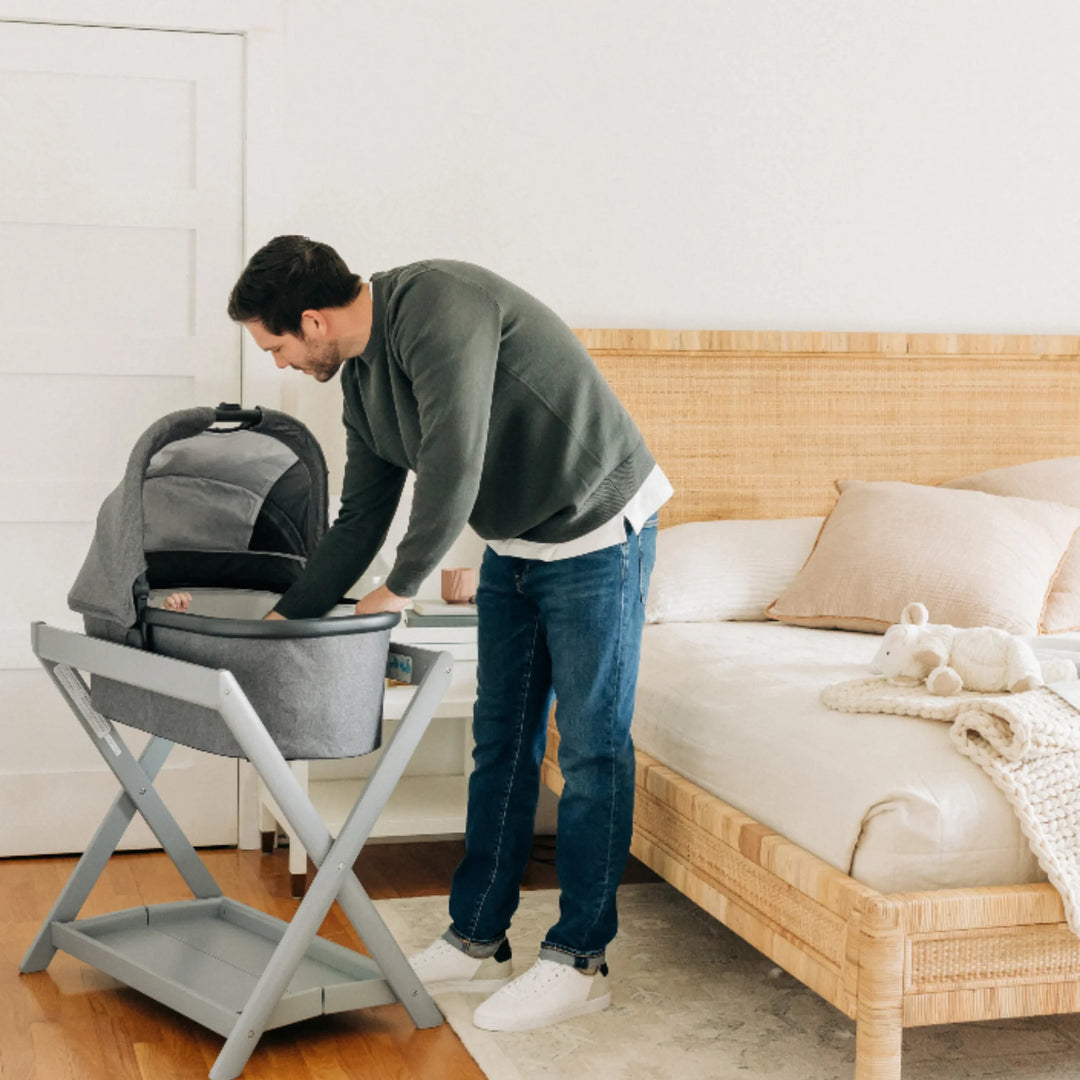 Parent placing a baby into the UPPAbaby Carrycot on the grey UPPAbaby Carrycot Stand beside a bed, demonstrating the elevated safe-sleep setup in a modern bedroom.