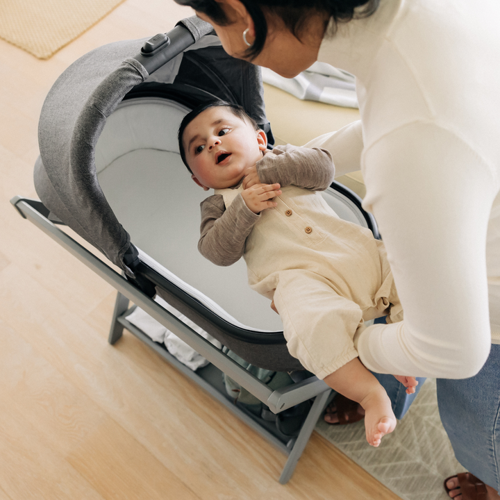 Mother putting her baby son to sleep in the carrycot that is on the carrycot stand. There are extra sleeping suits and the changing backpack on the convenient shelf on the stand under the carrycot.