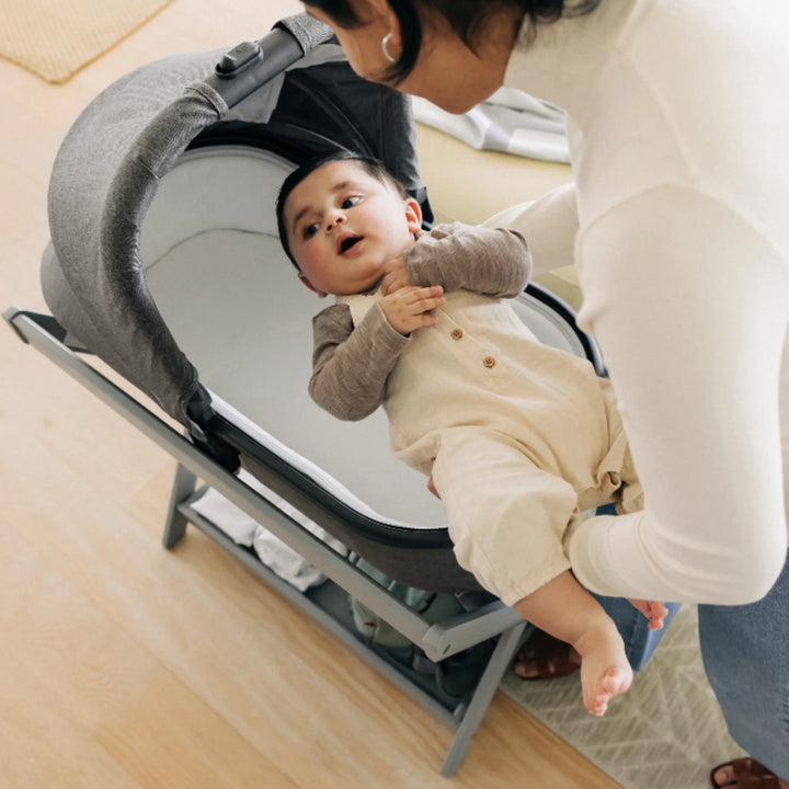Parent placing a baby into the UPPAbaby Carrycot on the matching stand, demonstrating the safe elevated sleep solution with a grey UPPAbaby carrycot in a bright modern home.