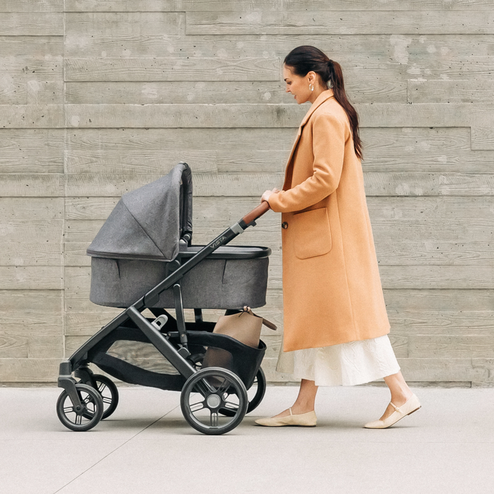 This image shows the Carrycot V3 in Greyson on the Vista V3 chassis whie a Mum in an orange coat walks with it.