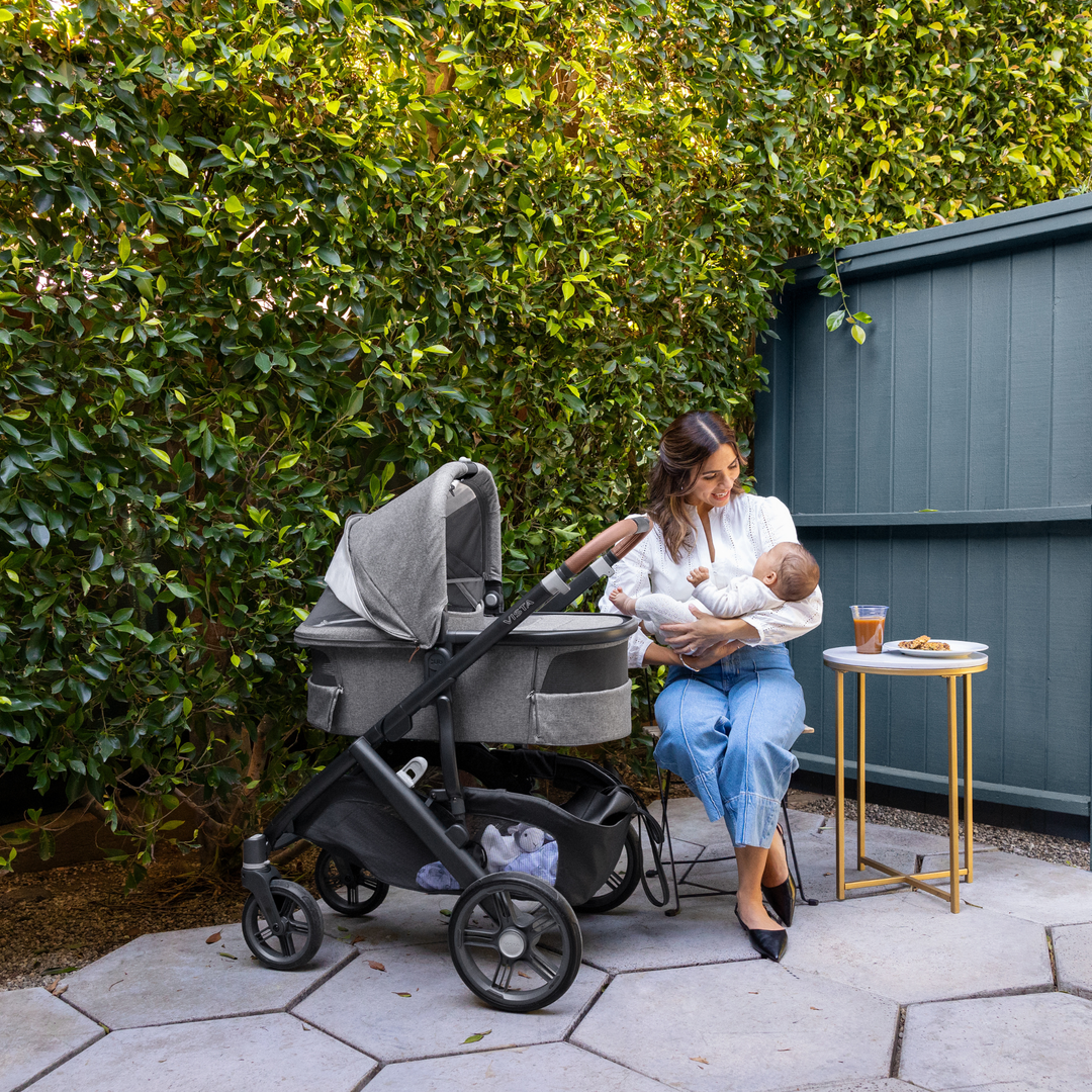 This image shows a Mum sitting outdoors enjoying a coffee and a pastry while holding her baby with the Carrycot V3 on the Vista V3 chassis with all of the vents open.