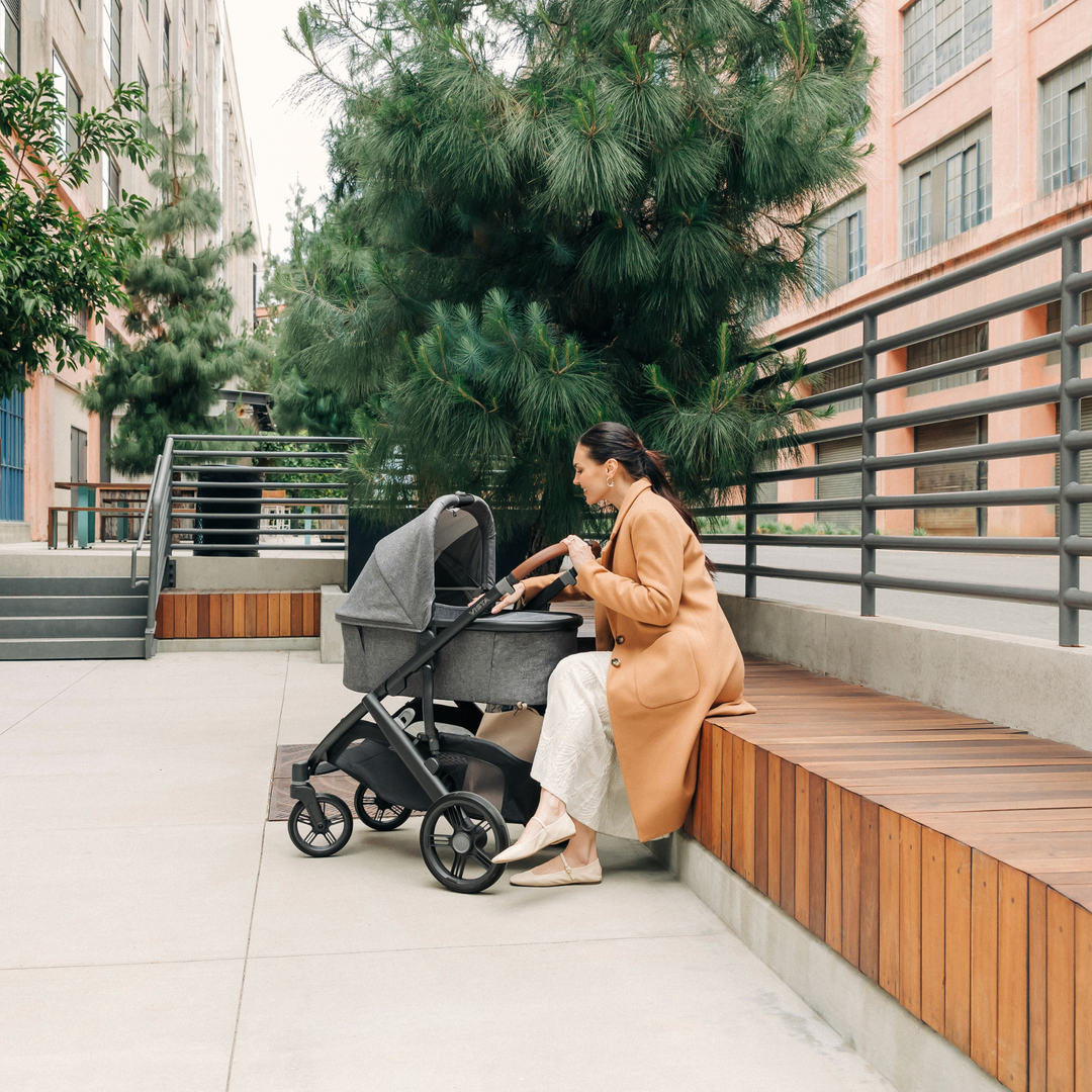 UPPAbaby Carrycot V3 on a stroller chassis while a Mum is sitting on a bench looking into the carrycot cooing at her baby.