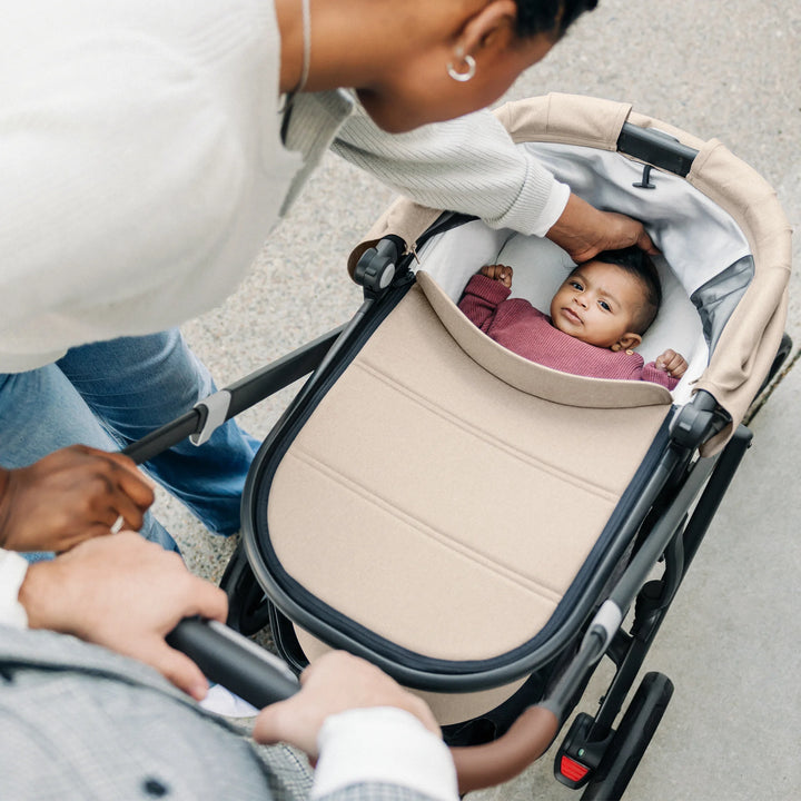 Baby lying comfortably in the UPPAbaby Vista V3 carrycot in the Liam oatmeal colourway, with caregivers attending overhead, showcasing the padded newborn bassinet, protective canopy and premium stroller design.
