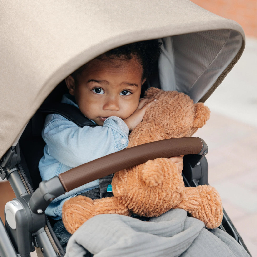 Baby seated in the UPPAbaby Vista V3 stroller in the Liam oatmeal colourway, cuddling a plush teddy under the extended canopy, highlighting the stroller’s padded seat, leather-accented bumper bar and premium sunshade design.