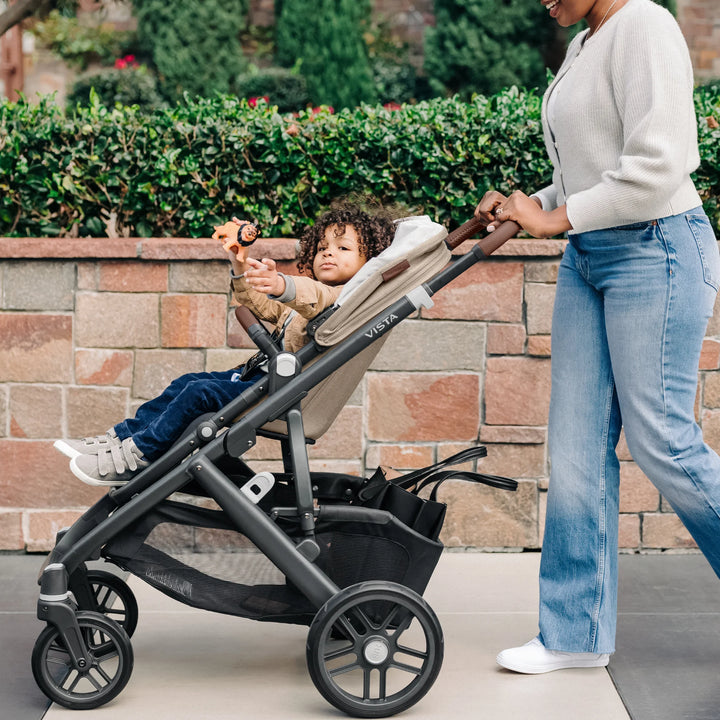 Parent pushing the UPPAbaby Vista V3 stroller in the Liam oatmeal colourway during an outdoor walk, with the toddler seated comfortably under the canopy, showcasing the stroller’s premium design, leather-accented handlebar, all-terrain wheels and large storage basket.