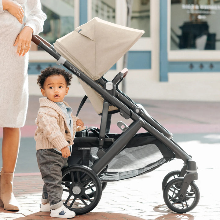 Toddler standing beside the UPPAbaby Vista V3 stroller in the Liam oatmeal colourway, showing the extended canopy, spacious under-seat storage basket, premium frame design and everyday use in an urban setting.