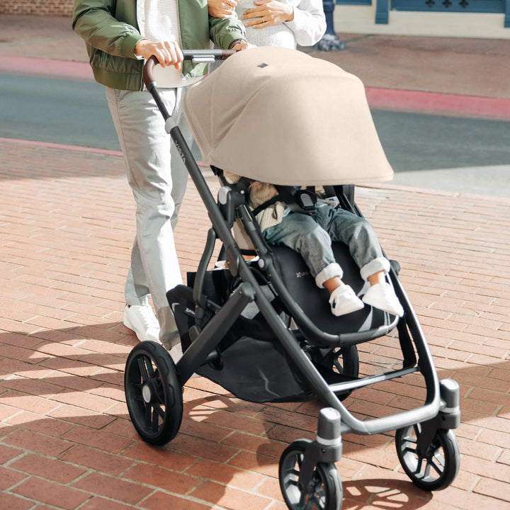 Parent pushing the UPPAbaby Vista V3 stroller in the Liam oatmeal colourway along a city walkway, with the toddler seated under the extended canopy, showcasing the stroller’s sleek design, all-terrain wheels and spacious under-seat basket.