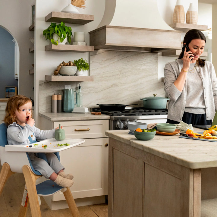 Woman talking on a phone in a kitchen with a child in a high chair.
