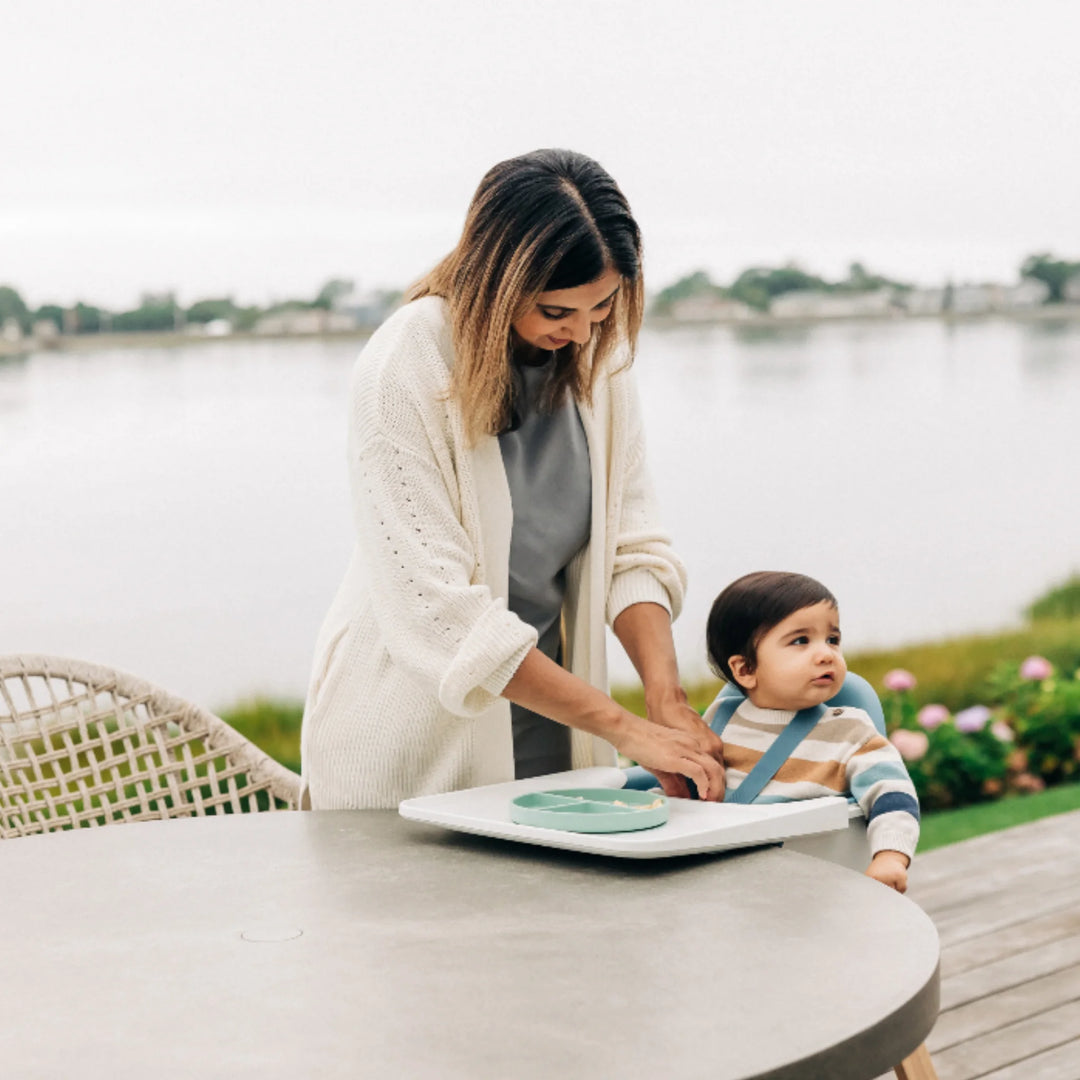 Mom helping toddler eat outdoors using UPPAbaby Ciro High Chair on patio overlooking lake, showcasing ergonomic baby seat and modern high-chair design for outdoor family dining.
