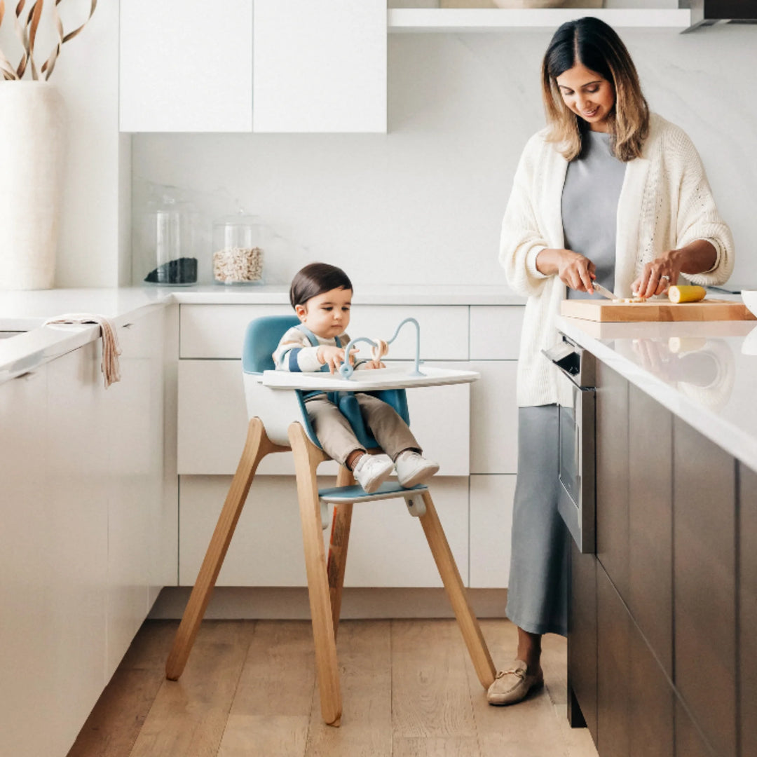 Mom preparing dinner in modern kitchen while toddler sits safely in UPPAbaby Ciro High Chair, featuring ergonomic design, adjustable tray and wooden legs.