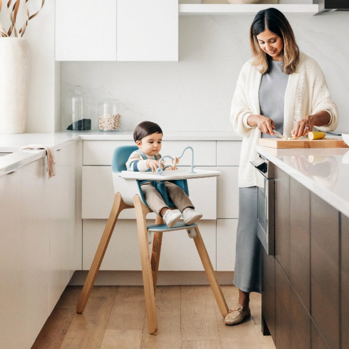 Mom preparing dinner in modern kitchen while toddler sits safely in UPPAbaby Ciro High Chair, featuring ergonomic design, adjustable tray and wooden legs.