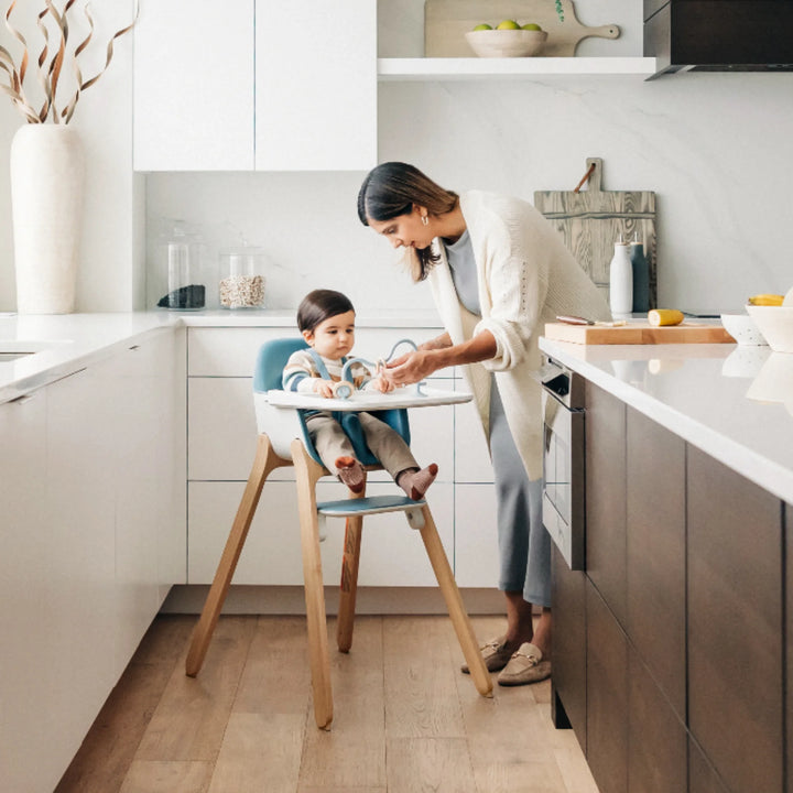 Mother feeding toddler seated in UPPAbaby Ciro High Chair in modern kitchen, showcasing ergonomic design, adjustable tray and stylish wooden legs.