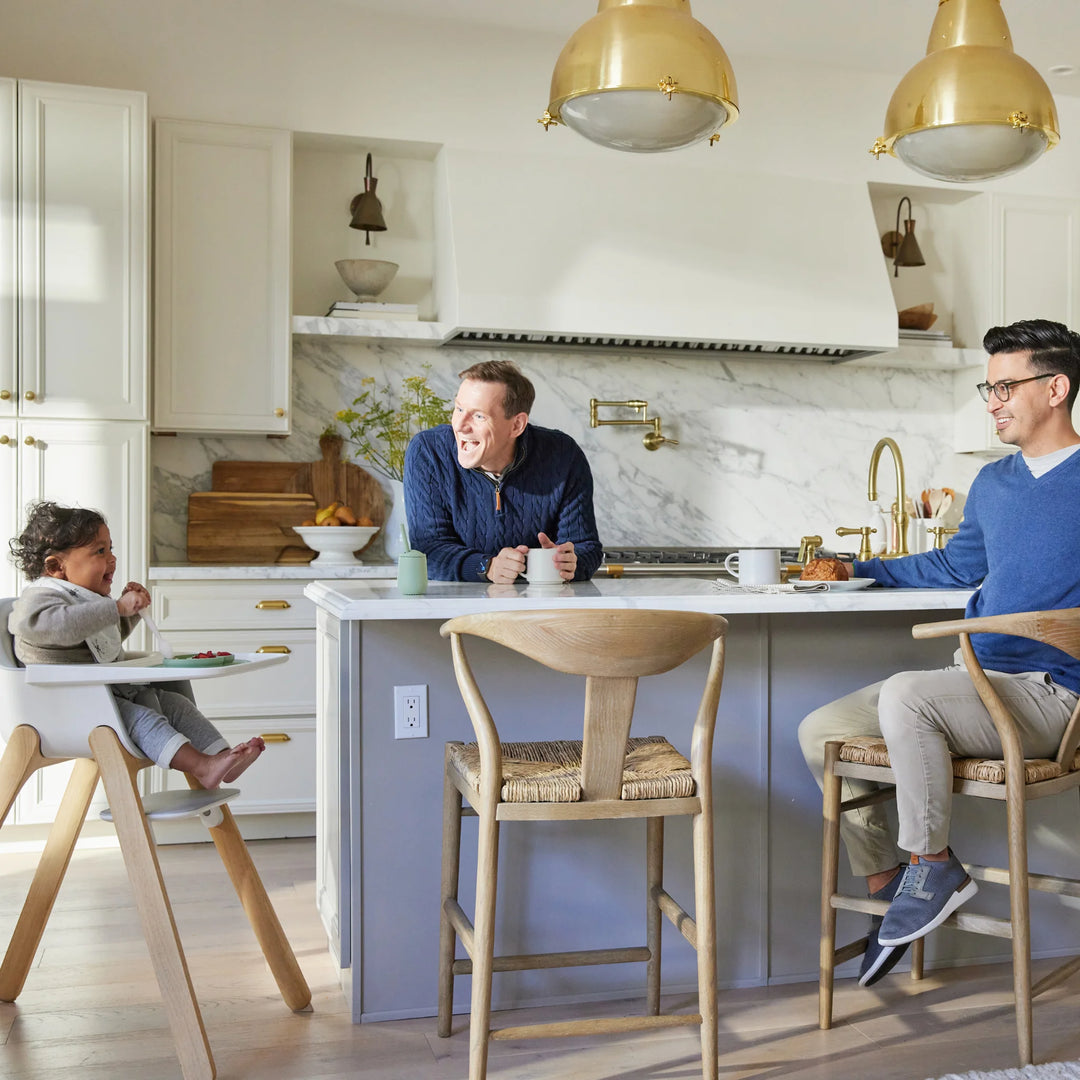 Family in a modern kitchen with marble backsplash and gold pendant lights with child laughing in grey modern high chair.