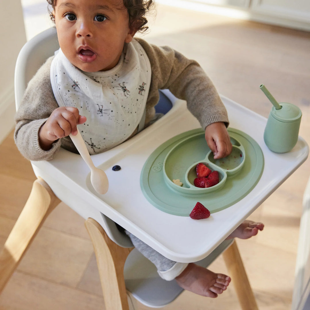Child in a high chair with a tray, spoon, and cup, sitting on a wooden floor.