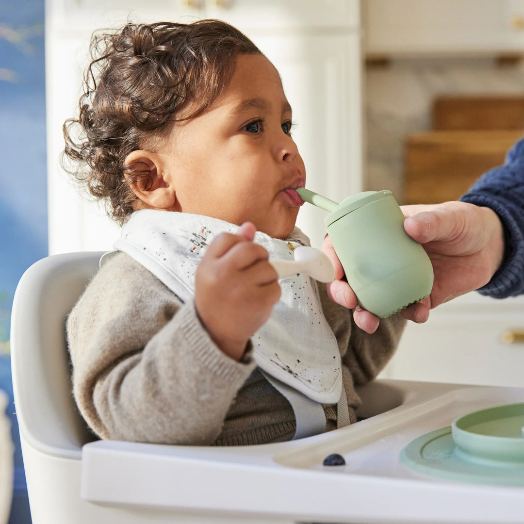 Child in a high chair being fed with a green sippy cup by an adult