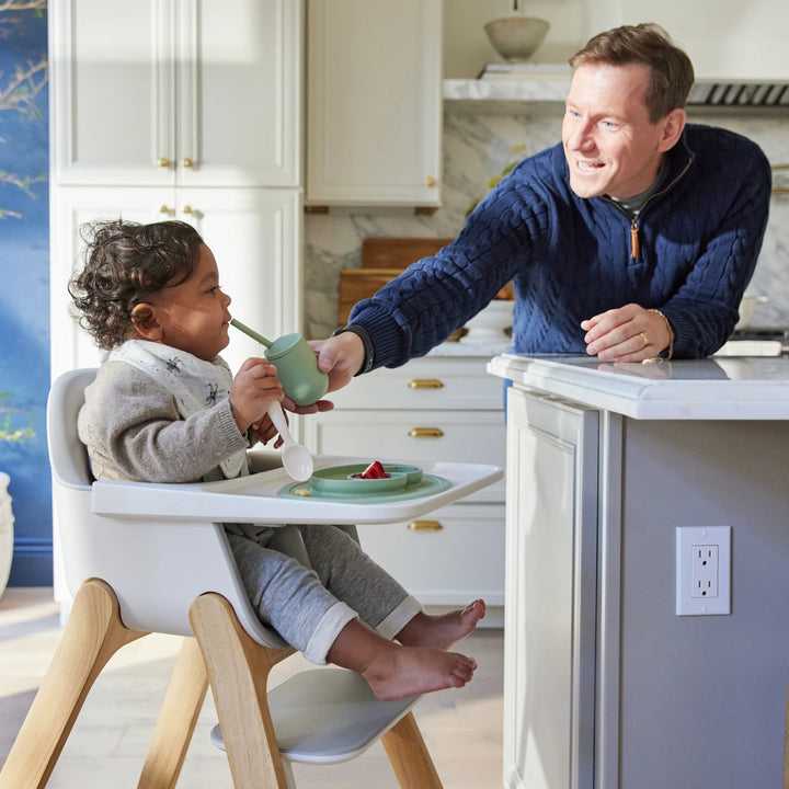 Man feeding a child in a grey high chair in a modern kitchen.