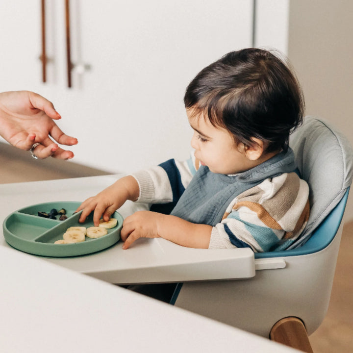 Toddler seated in an UPPAbaby Ciro high chair with grey cushion accessory, eating from a divided silicone plate while an adult hand offers food during mealtime.