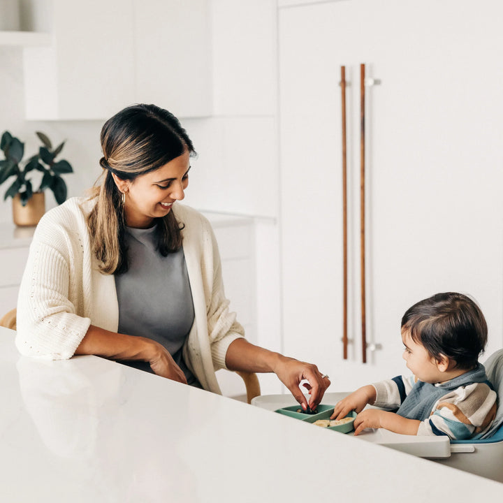 Smiling adult feeding a toddler seated in an UPPAbaby Ciro high chair with grey cushion accessory at a bright kitchen counter during mealtime