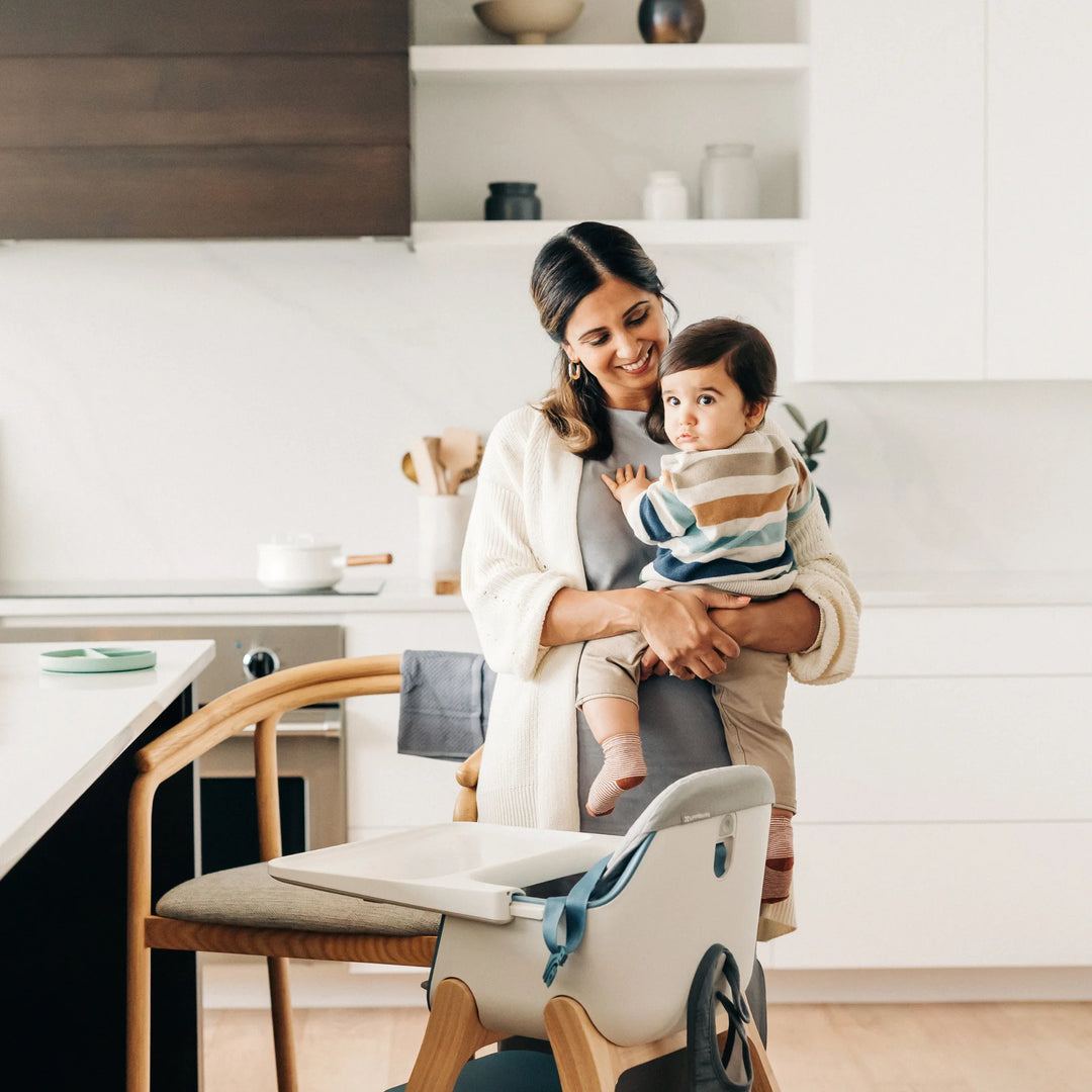 Adult holding a toddler beside an UPPAbaby Ciro high chair with grey cushion accessory in a bright modern kitchen after mealtime.