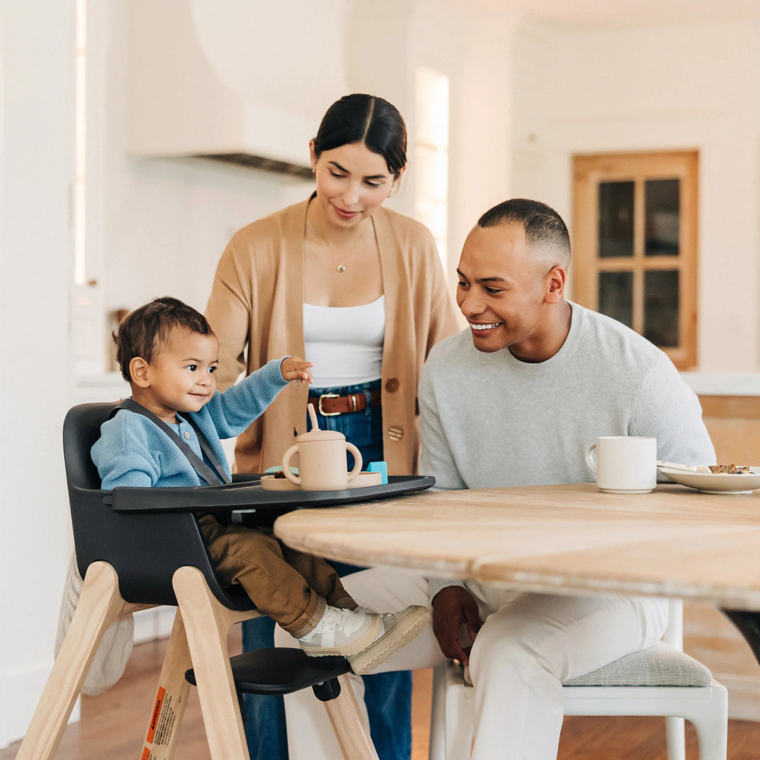 Family of three sitting at a kitchen table with a child in a black high chair.