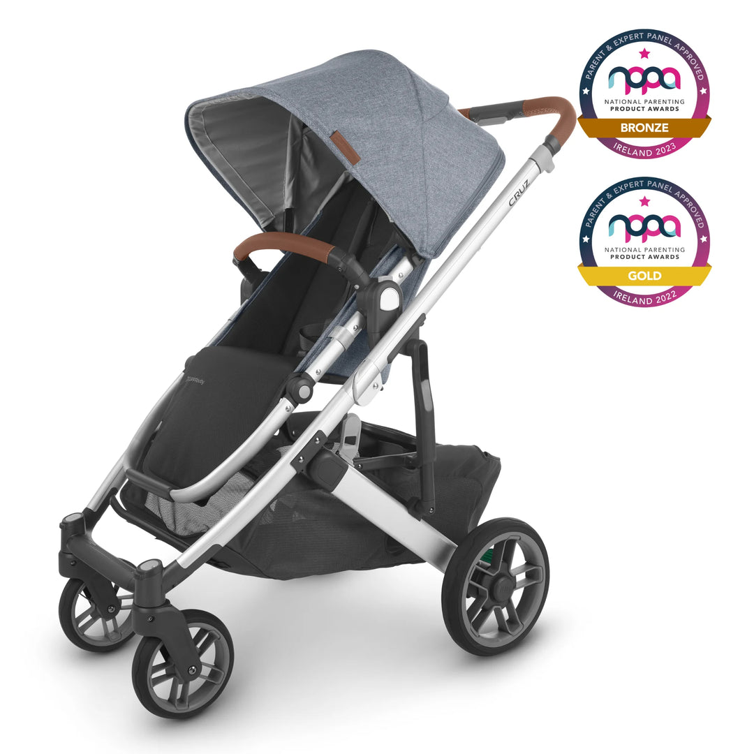 The UPPAbaby Cruz V2 in Gregory has a blue melange fabric, silver frame and saddle REACH certified leather accents. The stroller is angled to the left and two NPPA awards logos are visible in the top right of the stroller