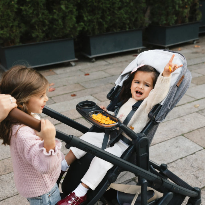 Child riding in UPPAbaby Cruz V2 stroller with Snack Tray, Greyson seat colour, while sibling stands beside stroller outdoors.