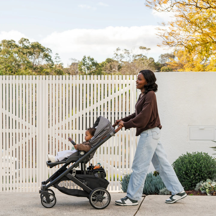 Smiling mum pushing child in Cruz V3 in Greyson in front of a white fence.