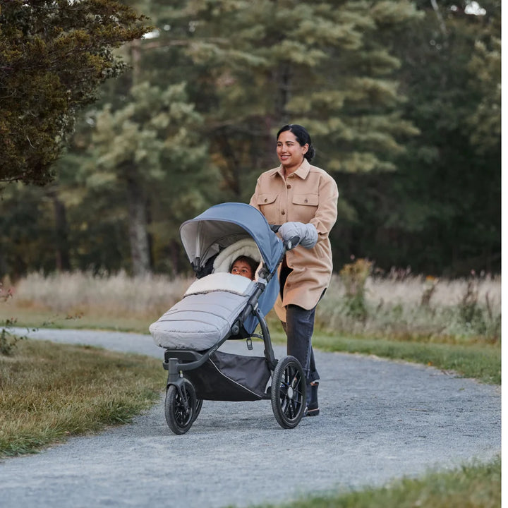 This image showcases a mum pushing her child along a gravel path in a wooded area on a cold day. The cozyganoosh footmuff is on the stroller to keep the child nice and warm.