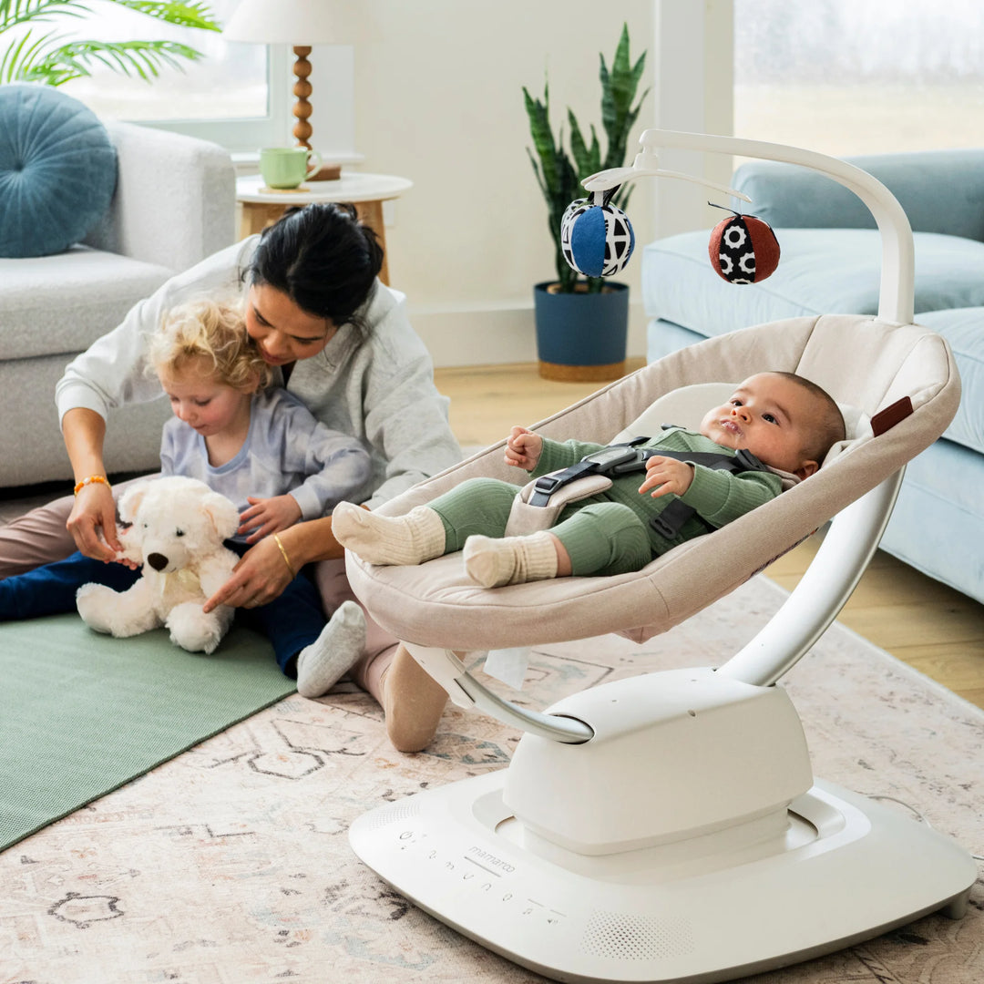 UPPAbaby MamaRoo smart swing (Charlie) in living room with baby reclining under toy mobile while parent plays with toddler nearby.