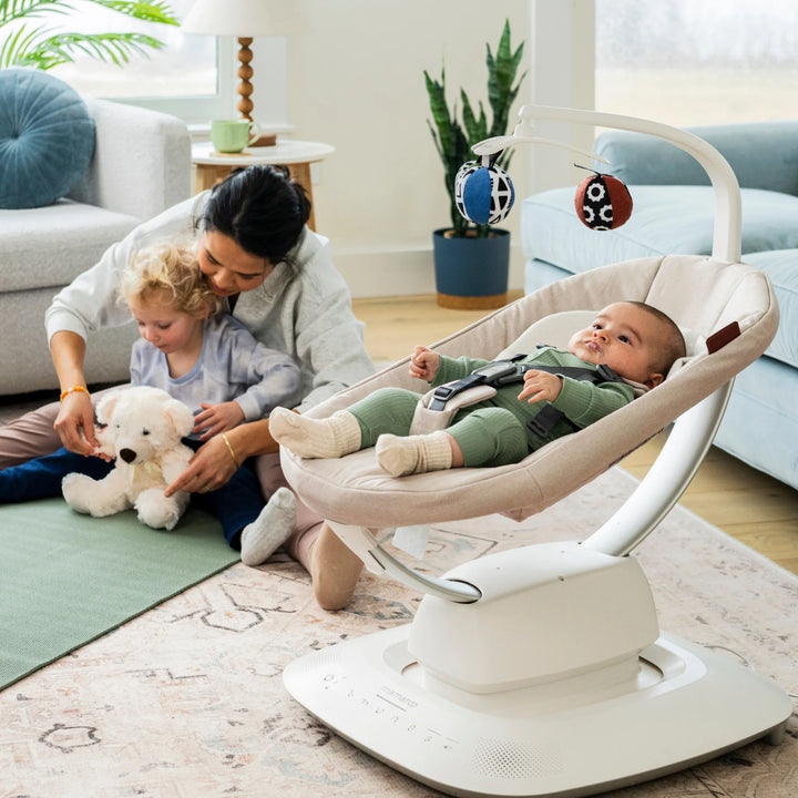UPPAbaby MamaRoo smart swing (Charlie) in living room with baby reclining under toy mobile while parent plays with toddler nearby.