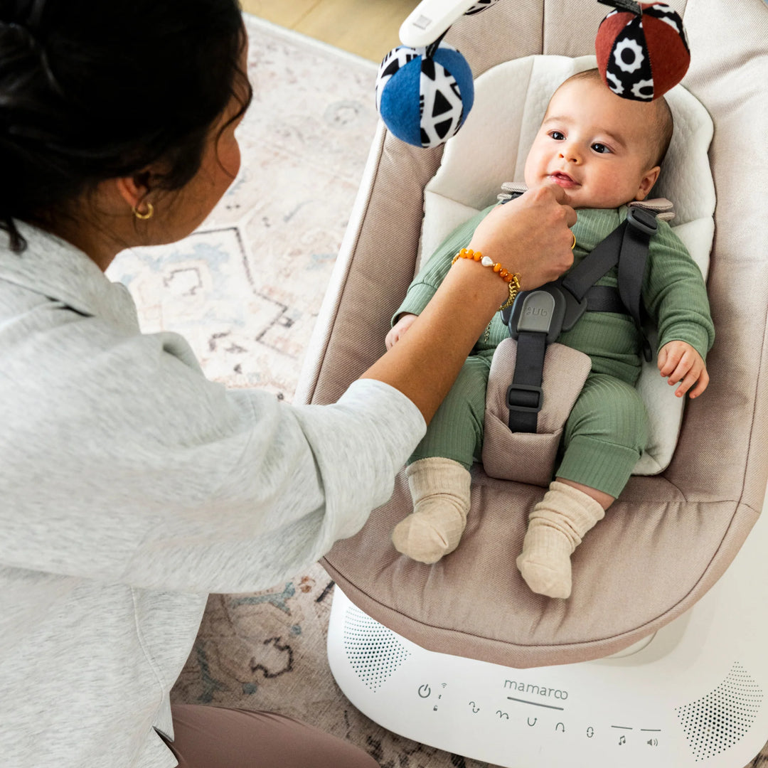 Baby relaxing in UPPAbaby MamaRoo smart swing (Charlie) with toy mobile while parent interacts at home.