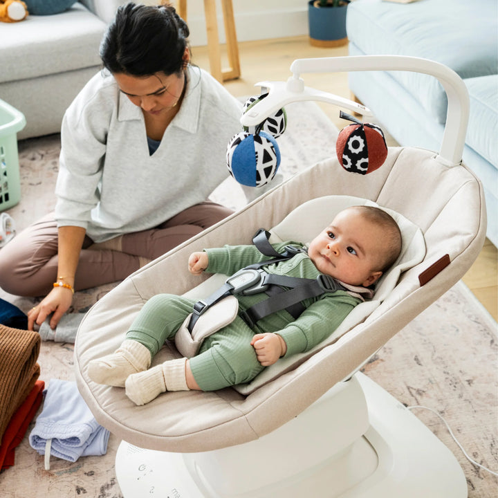 Baby relaxing in UPPAbaby MamaRoo smart swing (Charlie) with toy mobile while parent sits nearby at home.