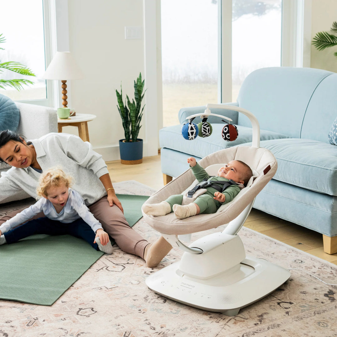 UPPAbaby MamaRoo smart swing (Charlie) in living room with baby reclining under toy mobile while parent stretches on yoga mat with toddler nearby.