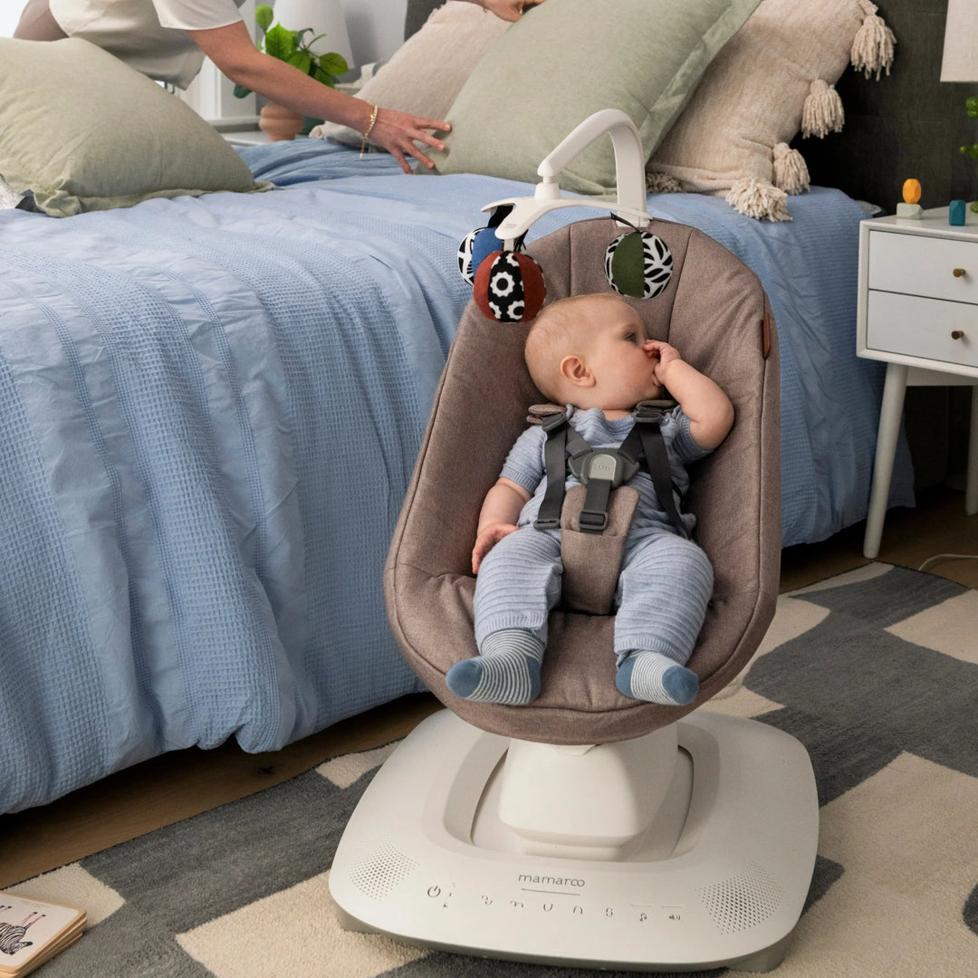 Baby sitting in the UPPAbaby MamaRoo Smart Swing in Wells fabric in a bedroom, with toy ball mobile overhead.