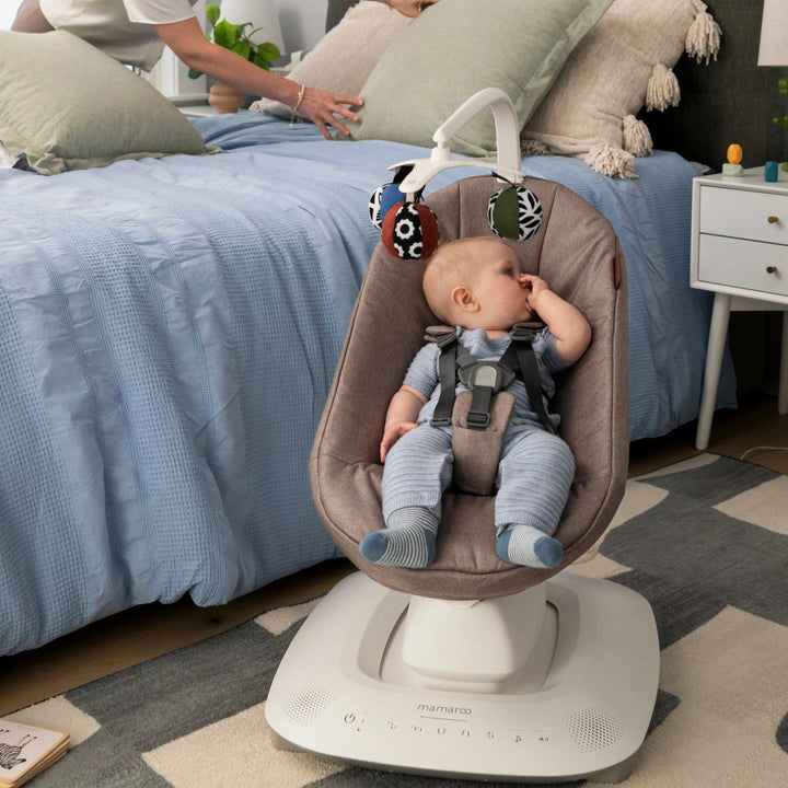 Baby sitting in the UPPAbaby MamaRoo Smart Swing in Wells fabric in a bedroom, with toy ball mobile overhead.