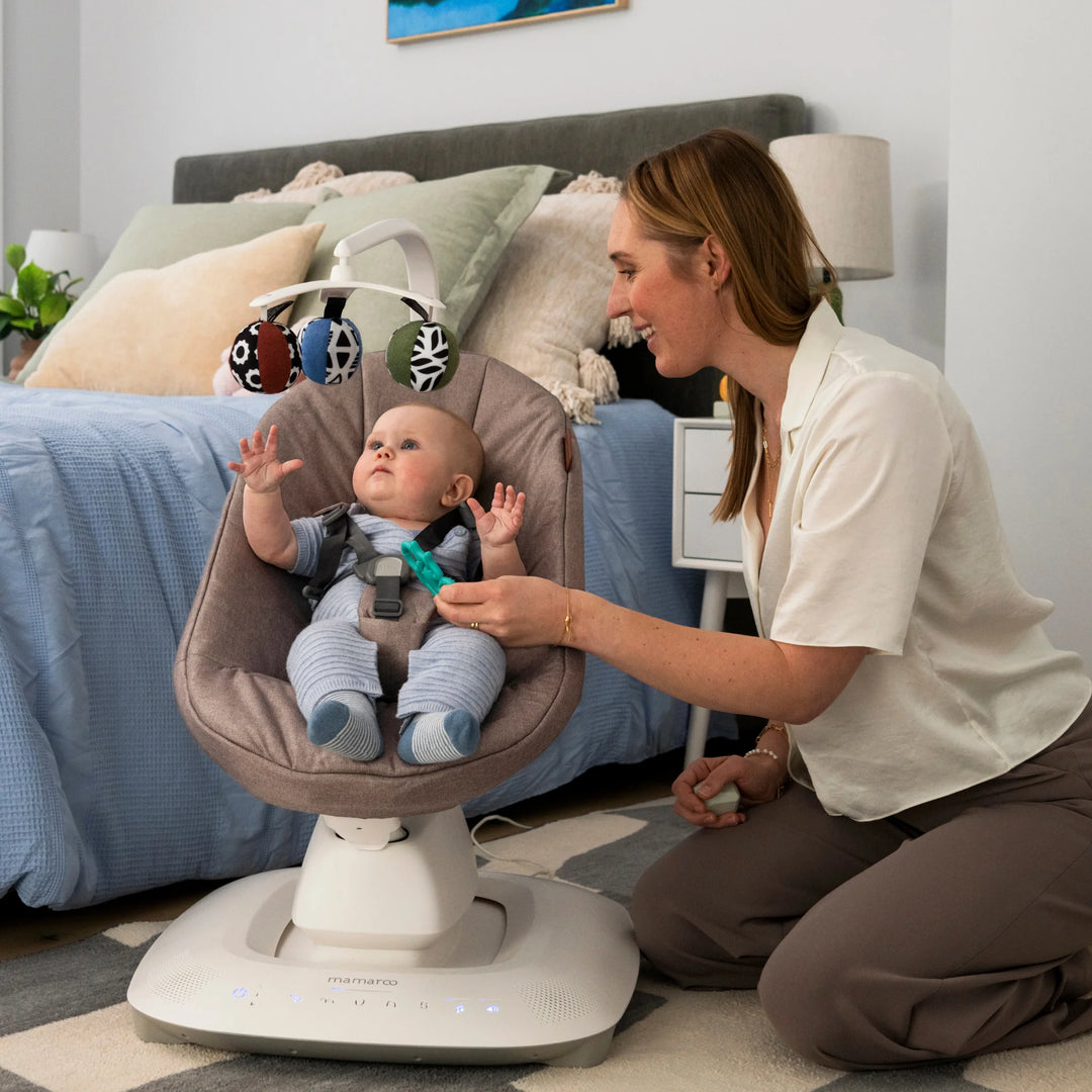 Baby relaxing in UPPAbaby MamaRoo smart swing (Wells) with toy mobile while parent interacts nearby in bedroom.