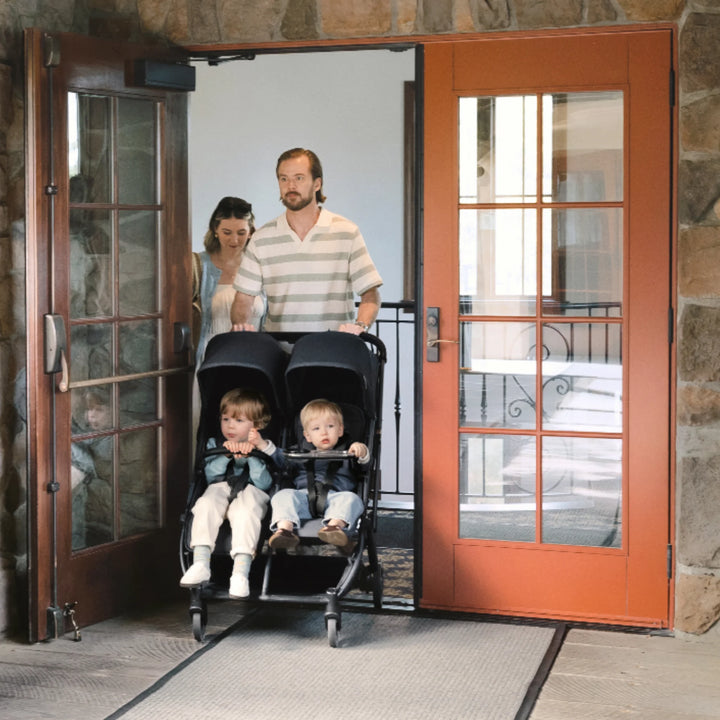 UPPAbaby Minu Duo Jake black double stroller fitting through a doorway, with two toddlers seated as parents push it outdoors