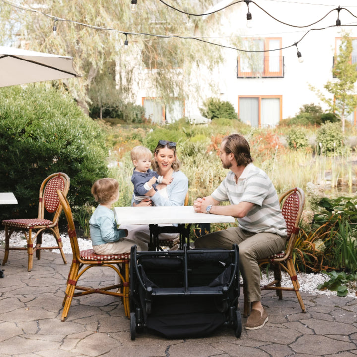 Folded UPPAbaby Minu Duo Jake black double stroller beside a family at an outdoor café table, compact fold for travel and storage