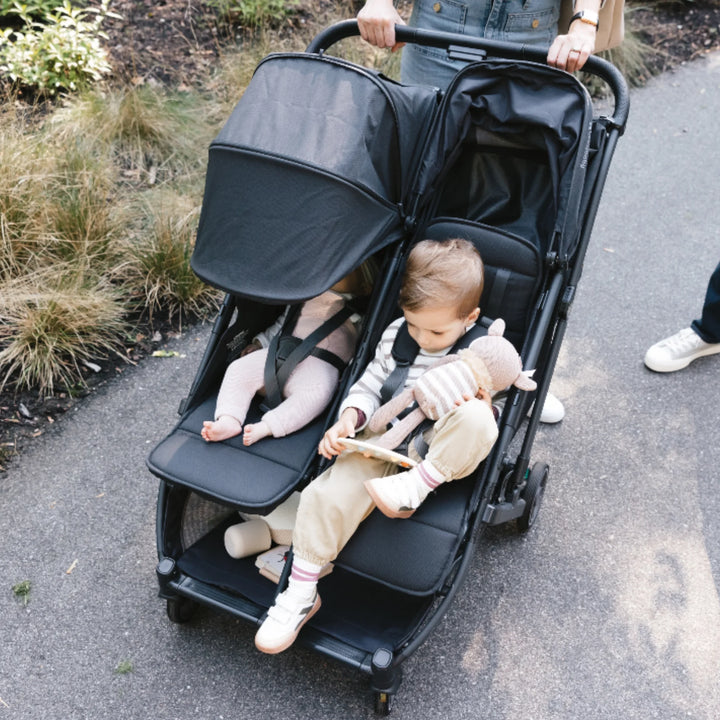 Two children riding in UPPAbaby Minu Duo double stroller in Jake black, compact twin/sibling pushchair on a park path