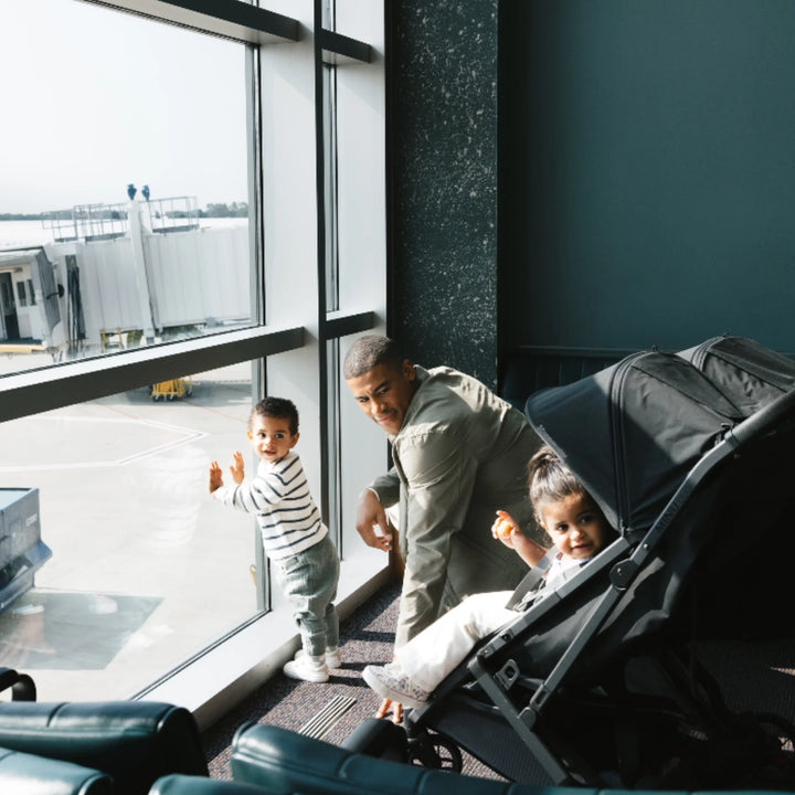 Family travelling with UPPAbaby Minu Duo Jake black double stroller at an airport gate, compact twin/sibling pushchair for travel