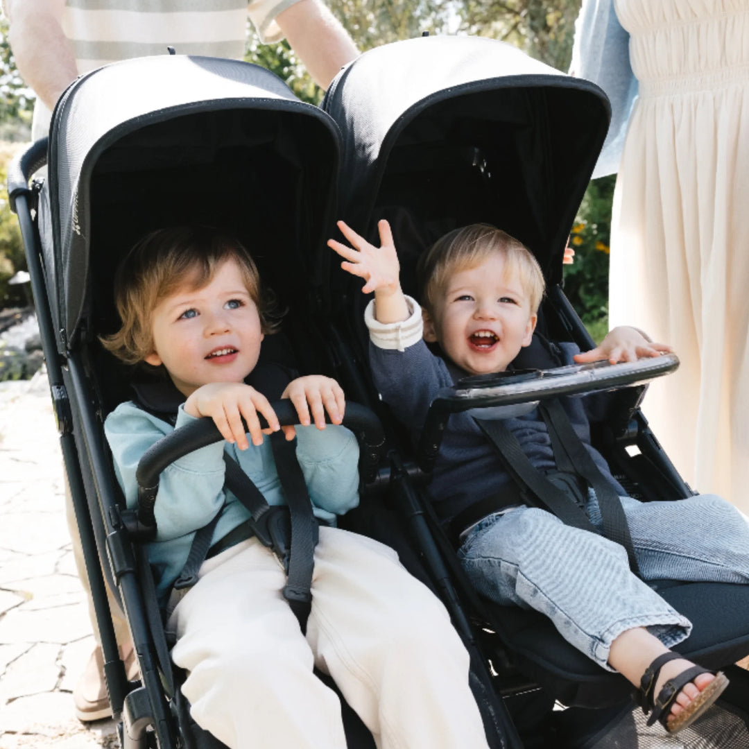 Two toddlers sitting in UPPAbaby Minu Duo double stroller using the snack tray accessory with cup holder, outdoors