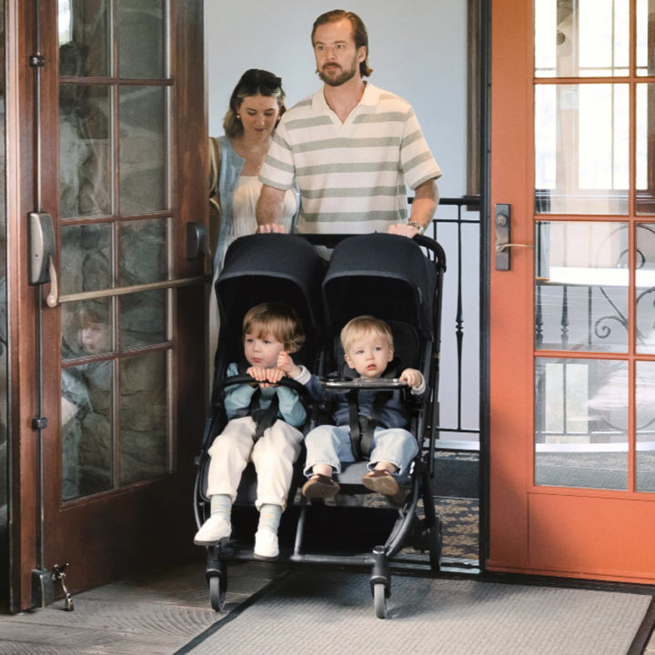 Two toddlers in UPPAbaby Minu Duo double stroller with snack tray accessory, being pushed through a doorway by parents