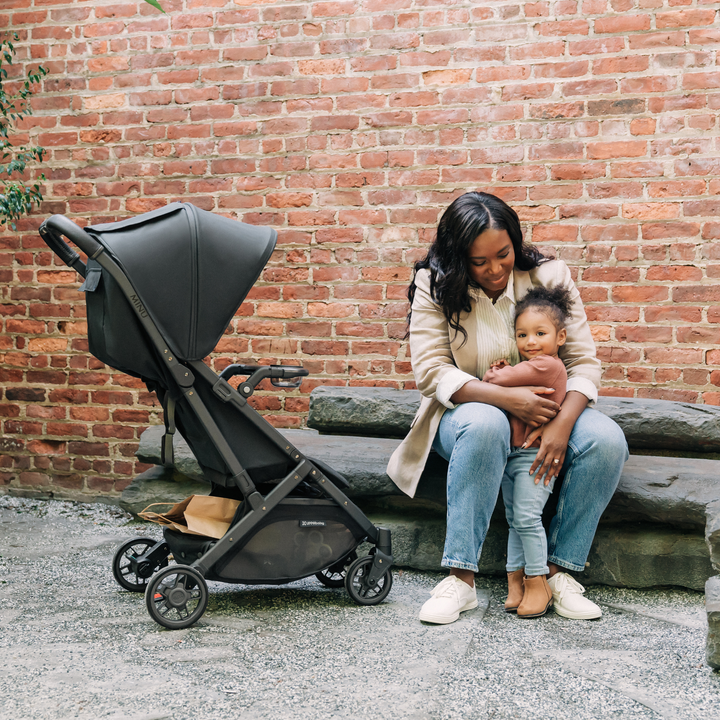 UPPAbaby Minu V2 in the Jake fashion with the Minu V2 Snack Tray attached. The stroller is angled towards a mother and her daughter happily sitting down on a stone slab bench together.