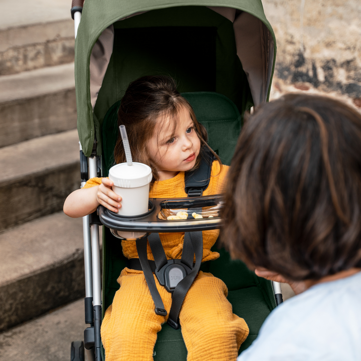 This is a lifestyle shot of the Minu V2 snack tray in use on the Minu V2 stroller in Emelia. A cute child is sitting in the pram and using the tray for crackers and a sippy cup