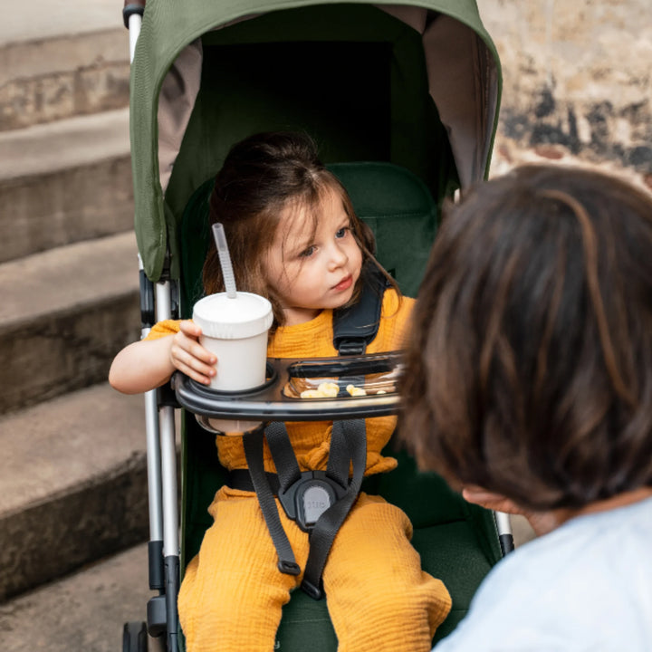 Toddler sitting in UPPAbaby Minu V2 travel stroller in Emilia with snack tray, holding a drink cup and snacks outdoors.