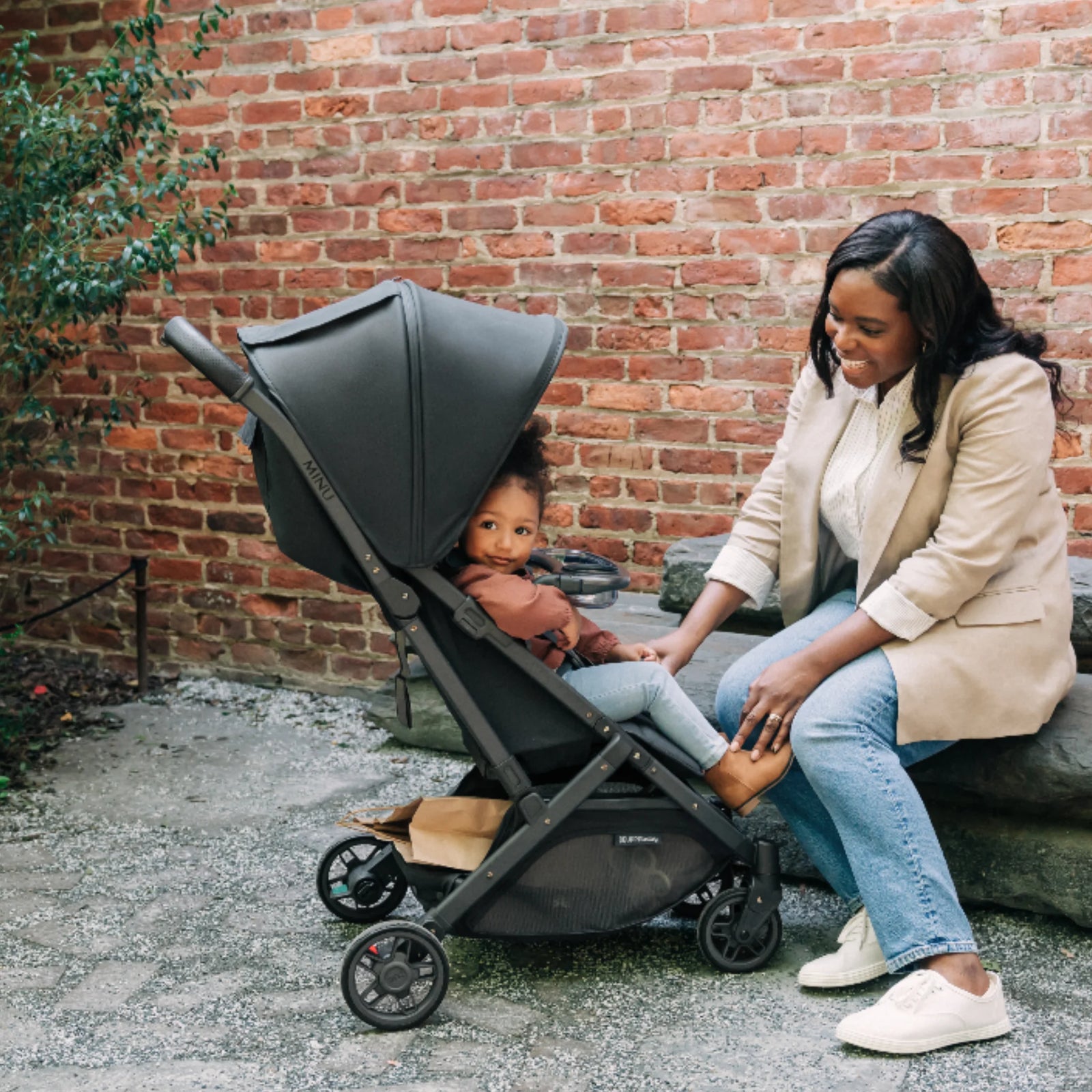 UPPAbaby Minu V2 compact stroller in Jake with snack tray accessory, shown with parent and child outdoors by a brick wall.