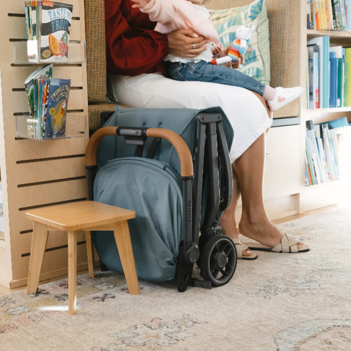 UPPAbaby MINU V3 stroller in Dillan blue folded beside seating in a library, compact travel stroller standing upright”