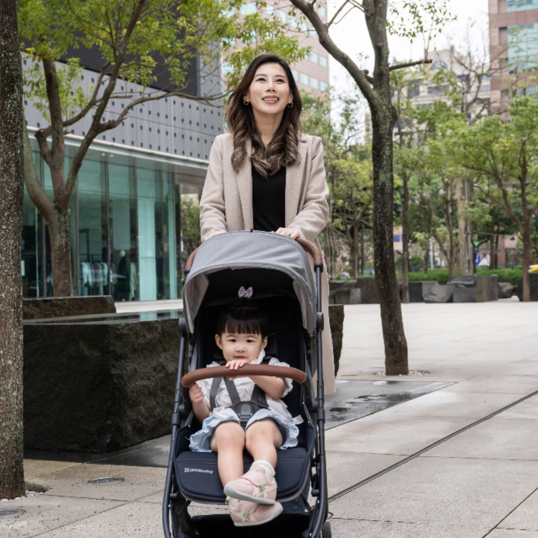 “Woman pushing the UPPAbaby MINU V3 stroller in Greysen with a toddler seated, walking through a tree-lined urban plaza.”